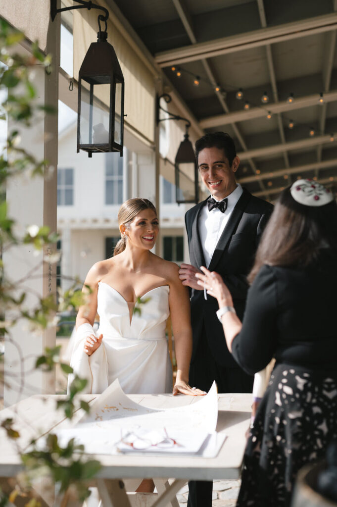 A bride and groom smiling and talking with a guest at a signing table, with the warm ambiance of a vineyard patio setting around them.