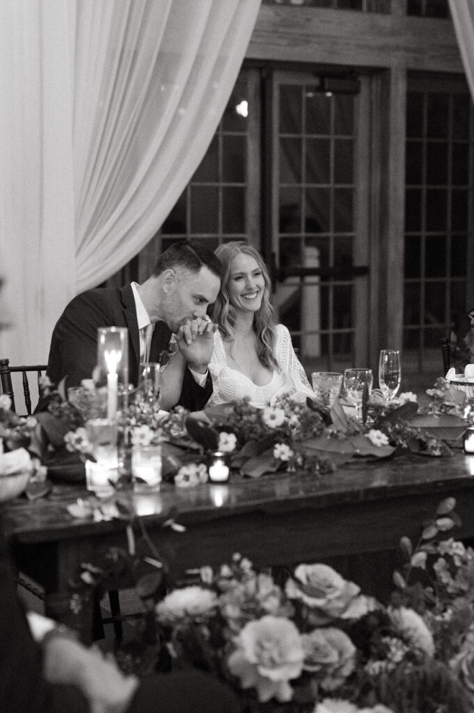 A monochrome image capturing an intimate moment at the wedding reception where the groom, dressed in a formal black suit, kisses the bride's hand, and both are smiling. The bride, in a white lace dress, is seated at a table adorned with candles and floral arrangements, contributing to a romantic and elegant atmosphere.