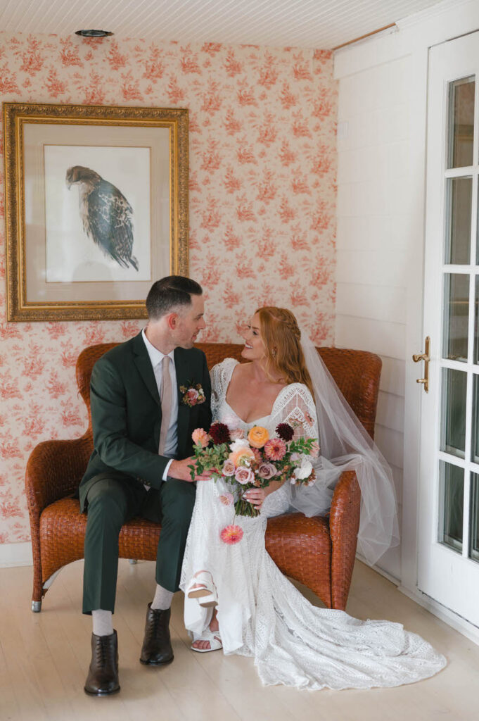 A bride and groom seated on an ornate wicker love seat, the bride in a white lace gown with a long veil and holding a bouquet of pastel flowers, and the groom in a deep green suit. They are looking into each other's eyes, with a backdrop of a wall covered in floral wallpaper and a framed image of a bird, radiating a vintage and romantic charm.