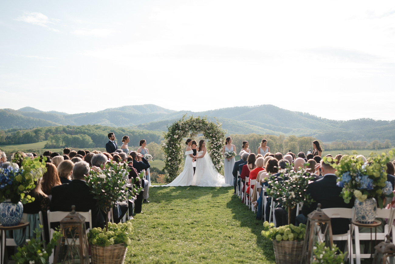 An outdoor wedding ceremony in progress with guests seated on either side of an aisle leading to a floral arch, set against a backdrop of rolling hills and a clear sky.