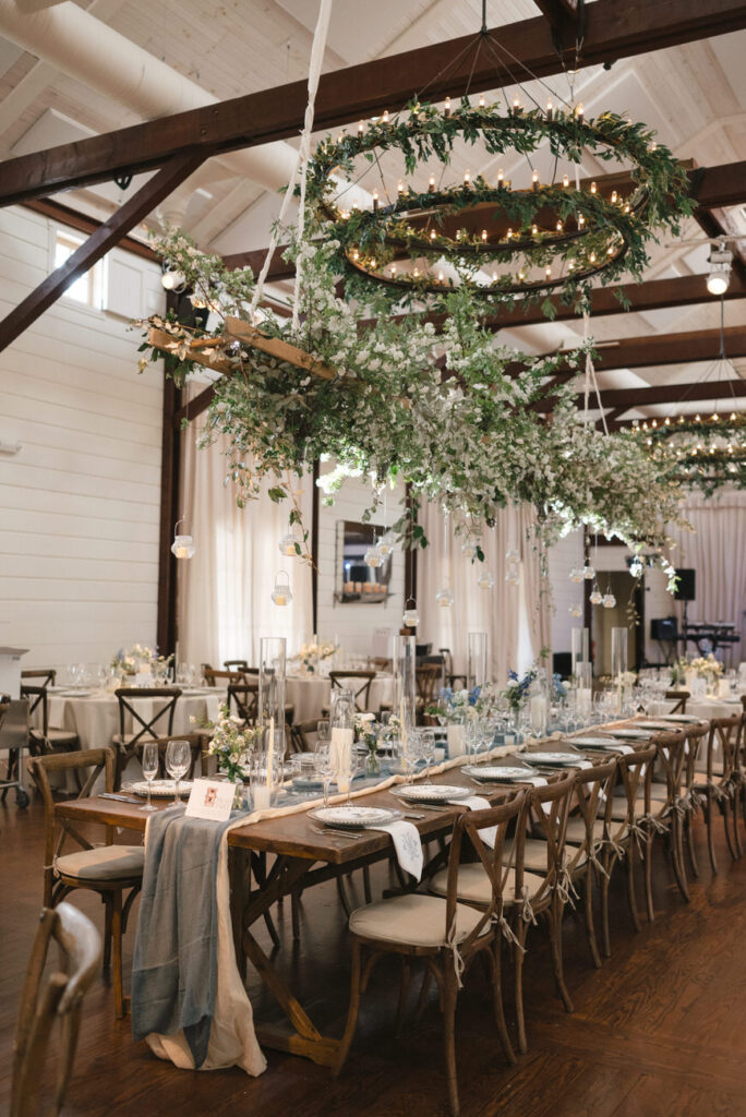 An elegant indoor wedding reception venue decorated with rustic wooden tables, adorned with lush floral centerpieces, and overhead circular lighting fixtures wrapped in greenery.