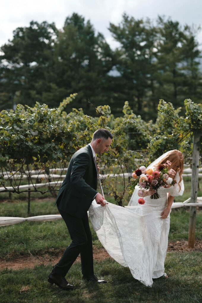 A bride and groom walking through a vineyard, the groom holding the bride's flowing lace dress, with a beautiful bouquet in her hand, amidst a backdrop of greenery.