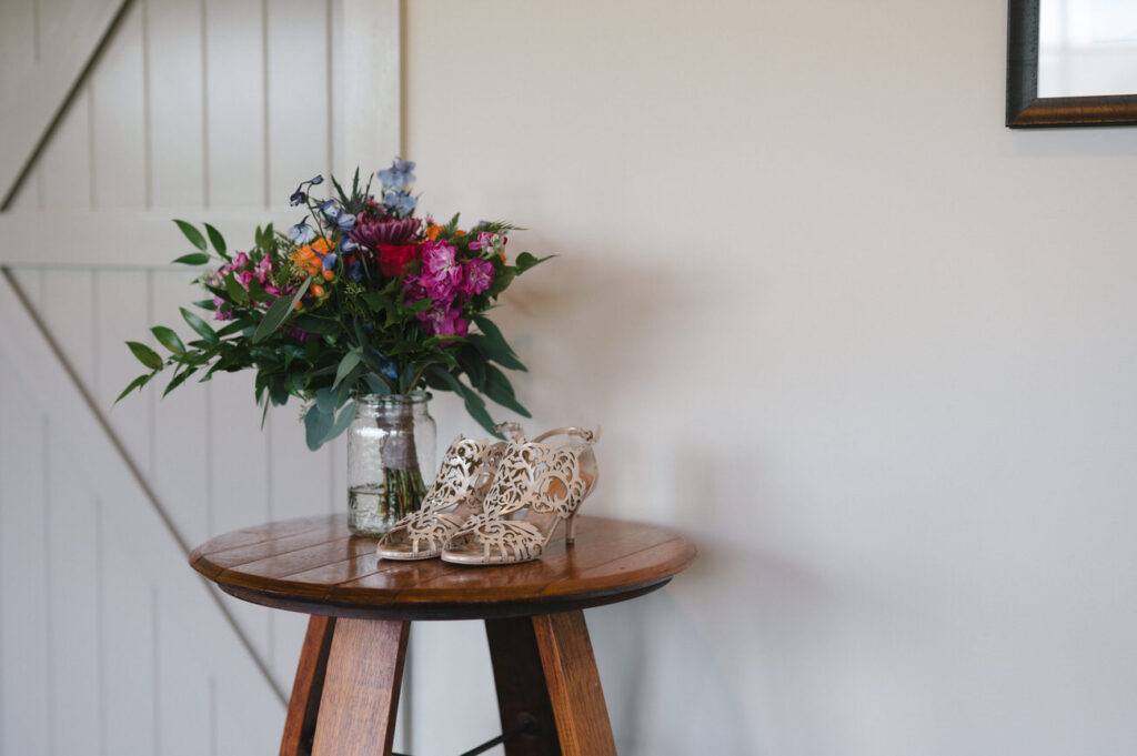 A side table with a vibrant floral arrangement in a mason jar next to a pair of ornate bridal shoes, creating a serene corner at a wedding venue.