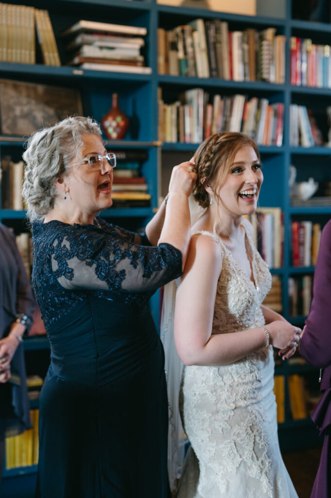 An older woman in a navy lace dress smiling as she adjusts a younger woman's hair, who is laughing joyfully in an embellished white wedding dress, against a backdrop of a bookshelf filled with books.