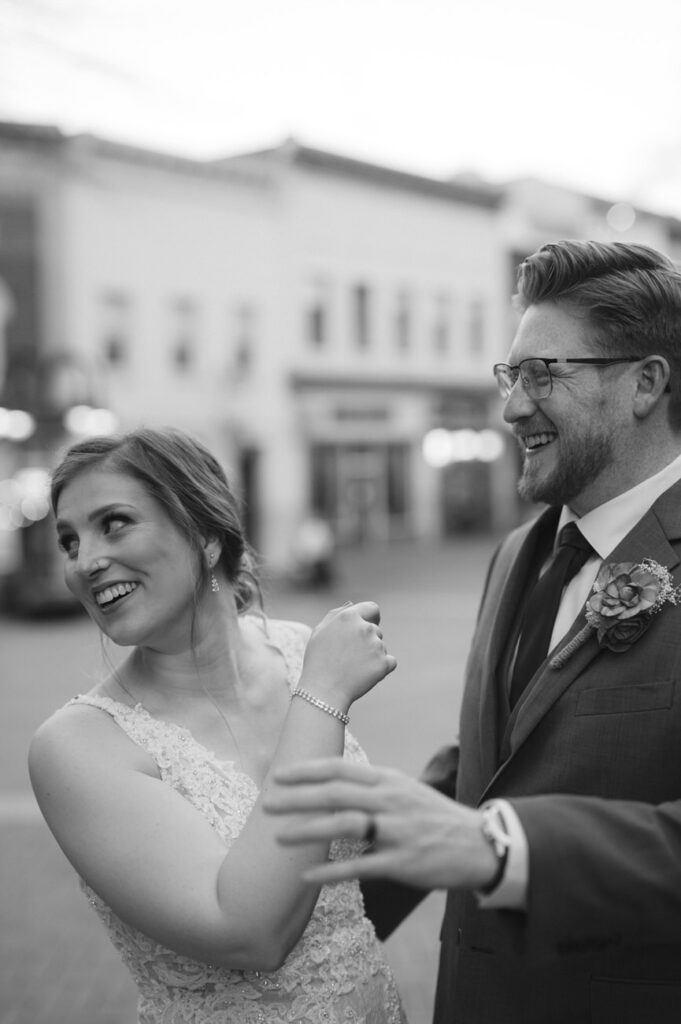 A candid black and white photo of a bride in a lace dress, smiling and dancing with a man in a smart suit and glasses, on a city street.