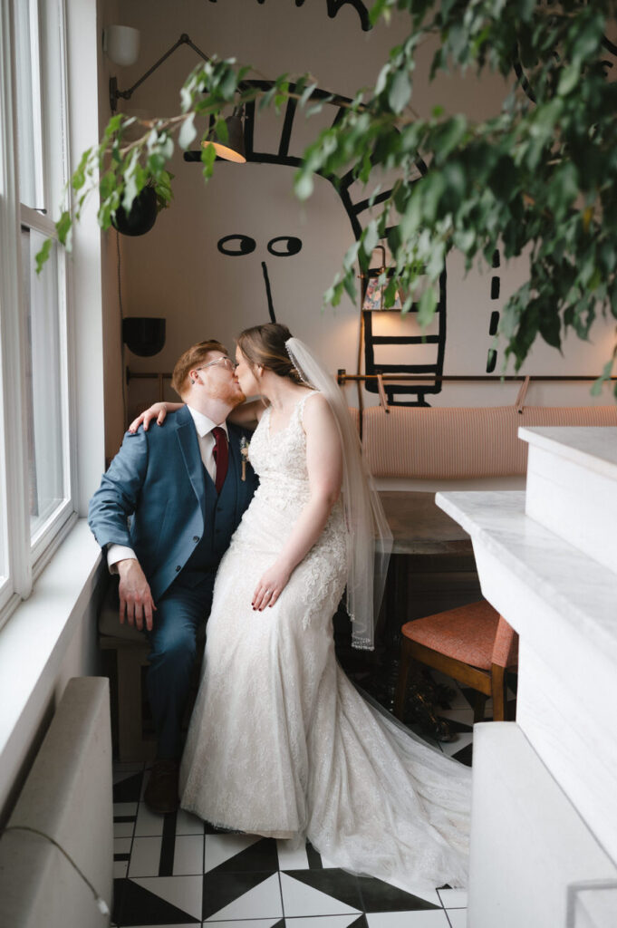 A bride and groom sharing a romantic kiss, seated by a window with a whimsical black and white wall art in the background, illustrating a cozy and intimate moment.