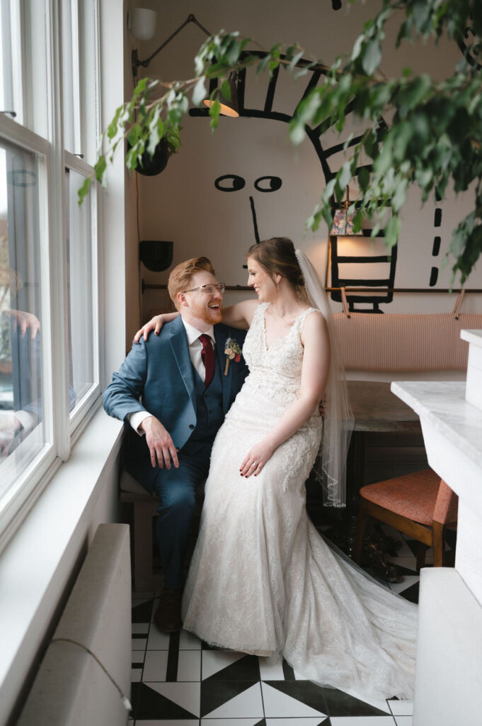 The same bride and groom smiling at each other, seated by the same window, with a relaxed and happy demeanor reflecting a candid and tender moment.
