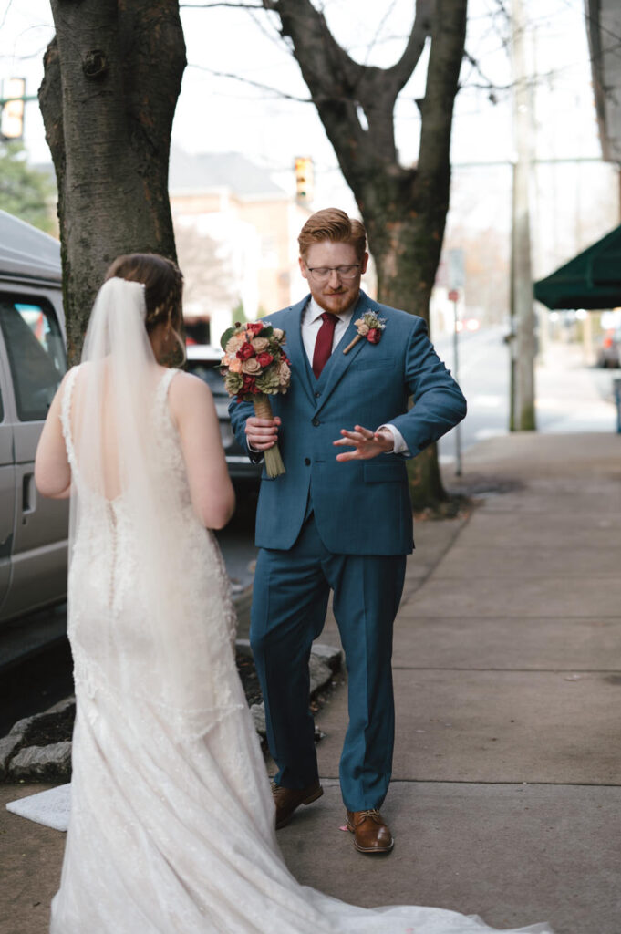 A groom in a blue suit gesturing animatedly, facing a bride with a cascading white veil and holding a bouquet with deep red flowers, creating a sense of playful conversation on a city sidewalk.