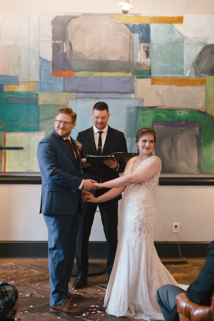 A bride and groom holding hands during the wedding ceremony, standing in front of an officiant with a colorful abstract painting in the background, symbolizing the vibrant start of their married life.