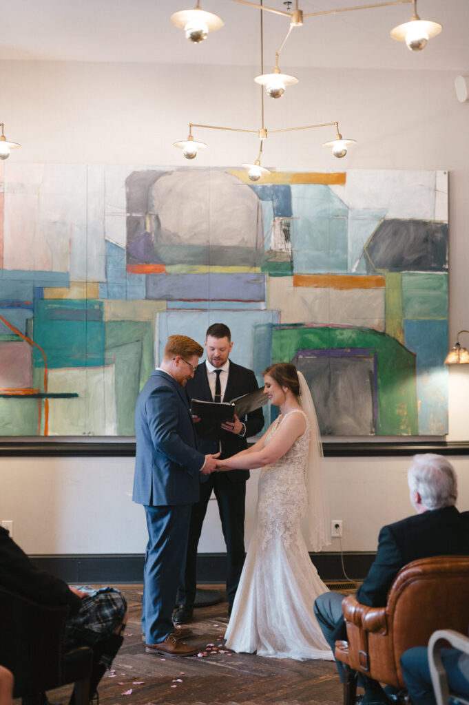 A bride and groom holding hands during the wedding ceremony, standing in front of an officiant with a colorful abstract painting in the background, symbolizing the vibrant start of their married life.