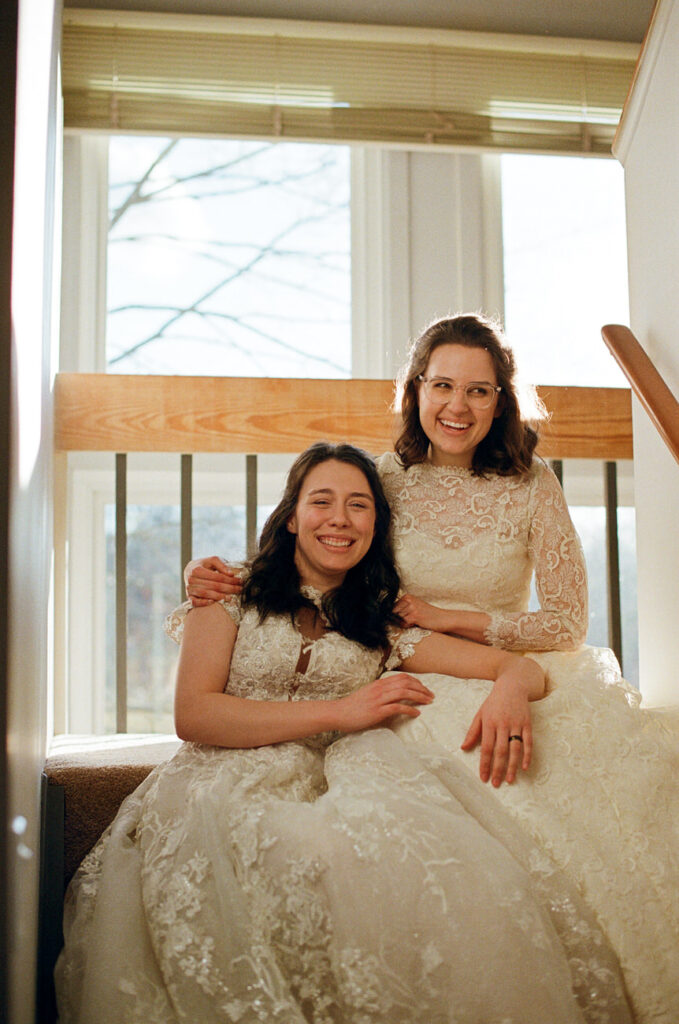 Two women in lace bridal gowns smiling while sitting in a sunlit room by a large window, with one embracing the other from behind.