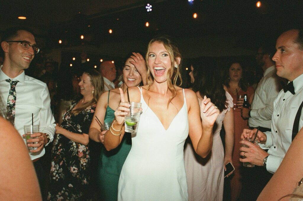 A lively wedding reception scene with guests dancing and a smiling woman in a sleeveless white dress holding a drink and dancing energetically.