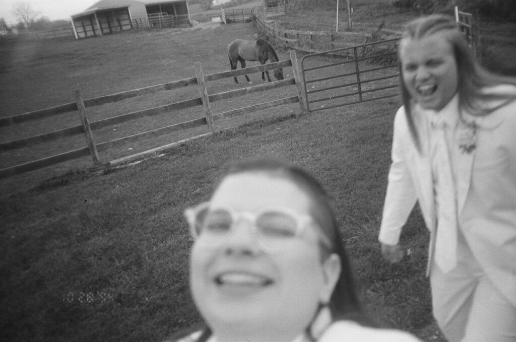 A black and white photo of two people, one in sharp focus with glasses, smiling in the foreground, and the other out of focus and laughing in the background, with a horse and a barn behind a fence visible in the distance.