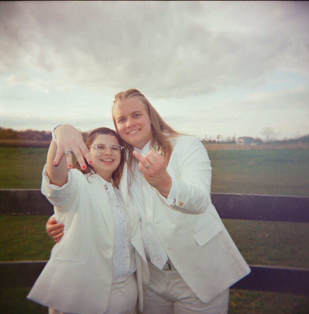 A color photograph of two individuals dressed in white suits, standing arm in arm behind a fence in an open field; both are showing their rings to the camera with smiles, under a cloudy sky.