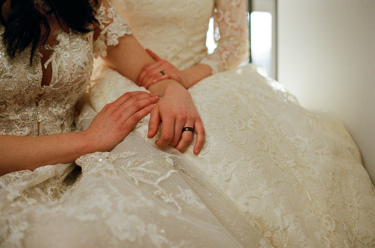 A close-up view highlighting the intricate lace detail of a wedding gown and the hands of two people, one with a wedding band, resting gently on the fabric.