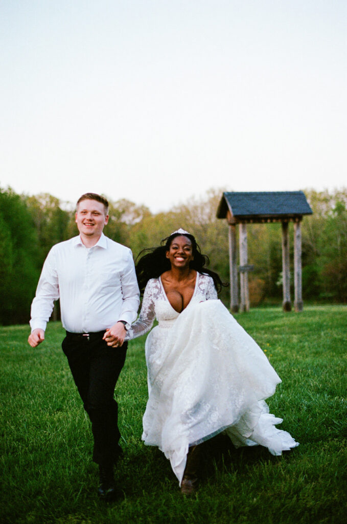 A joyous bride in a flowing wedding dress and a groom in a white shirt and black trousers running across a grassy field, with trees and a wooden structure in the background.