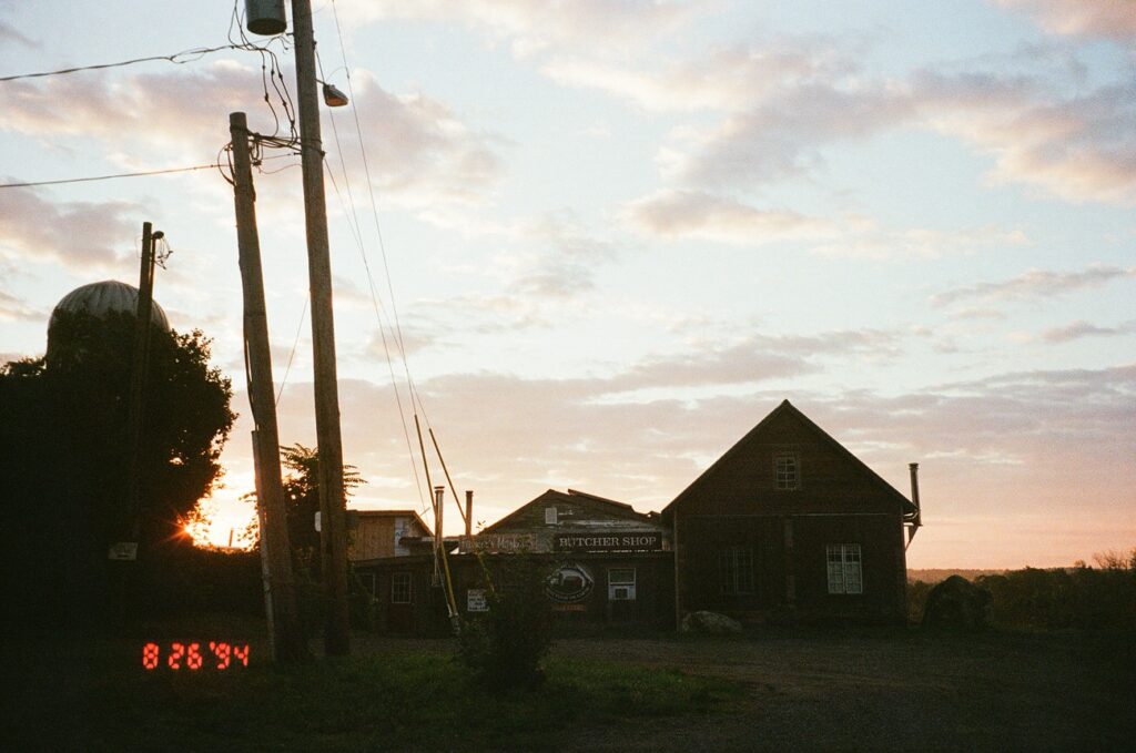A rural twilight scene featuring a building with a sign reading "Butcher Shop," with an electronic sign displaying the date '8 26 94' in red, under a sky with the setting sun.
