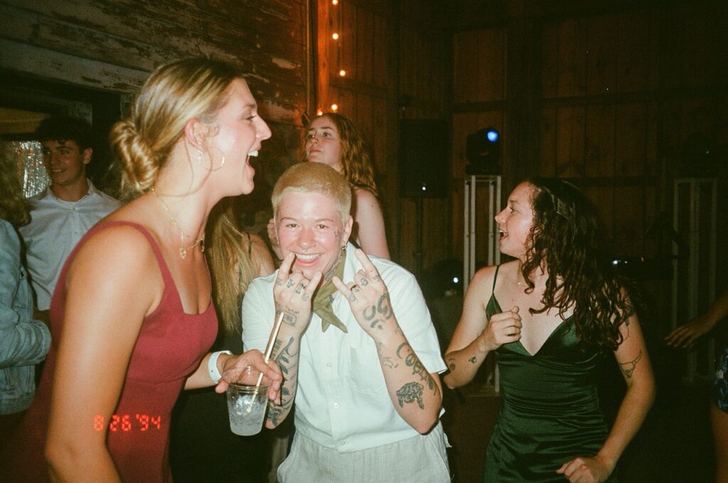 A vibrant indoor dance scene with three women, one with a bow tie and tattoos, laughing and enjoying the party, amidst a crowd.