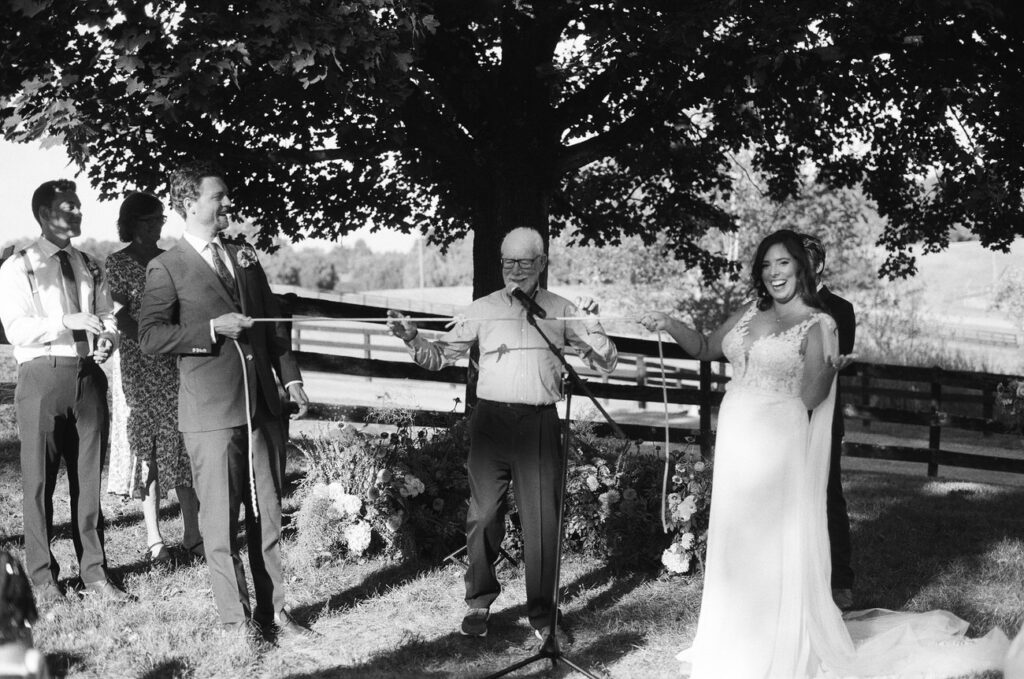 A black and white photo capturing a bride laughing during a speech at an outdoor ceremony, with guests and a tree in the background.