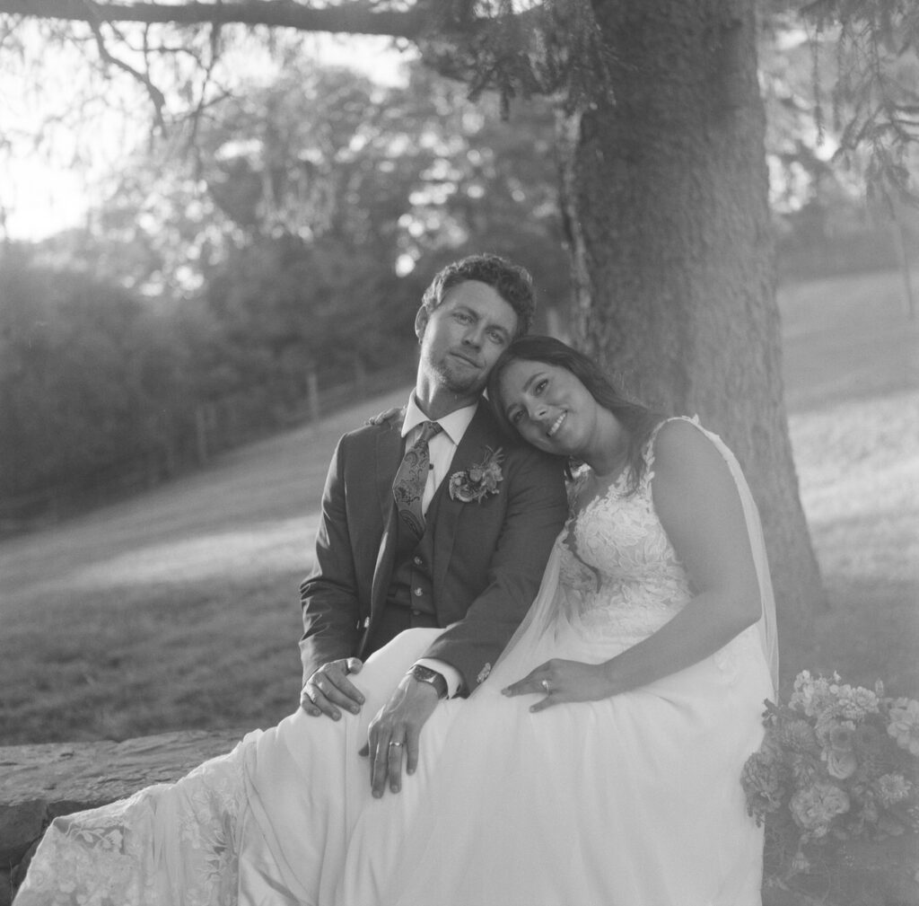 A serene black and white image of a bride and groom sitting under a large tree, the bride leaning on the groom with a tranquil expression.