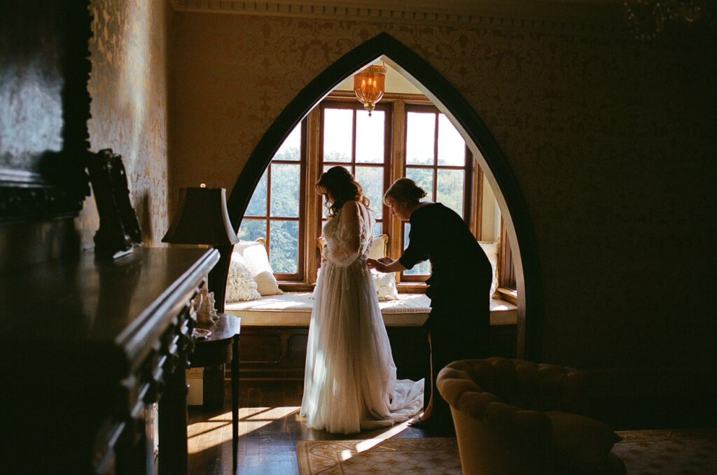 A bride in a long-sleeved wedding dress being assisted by another person in a dimly lit, elegantly furnished room with a large arched window.