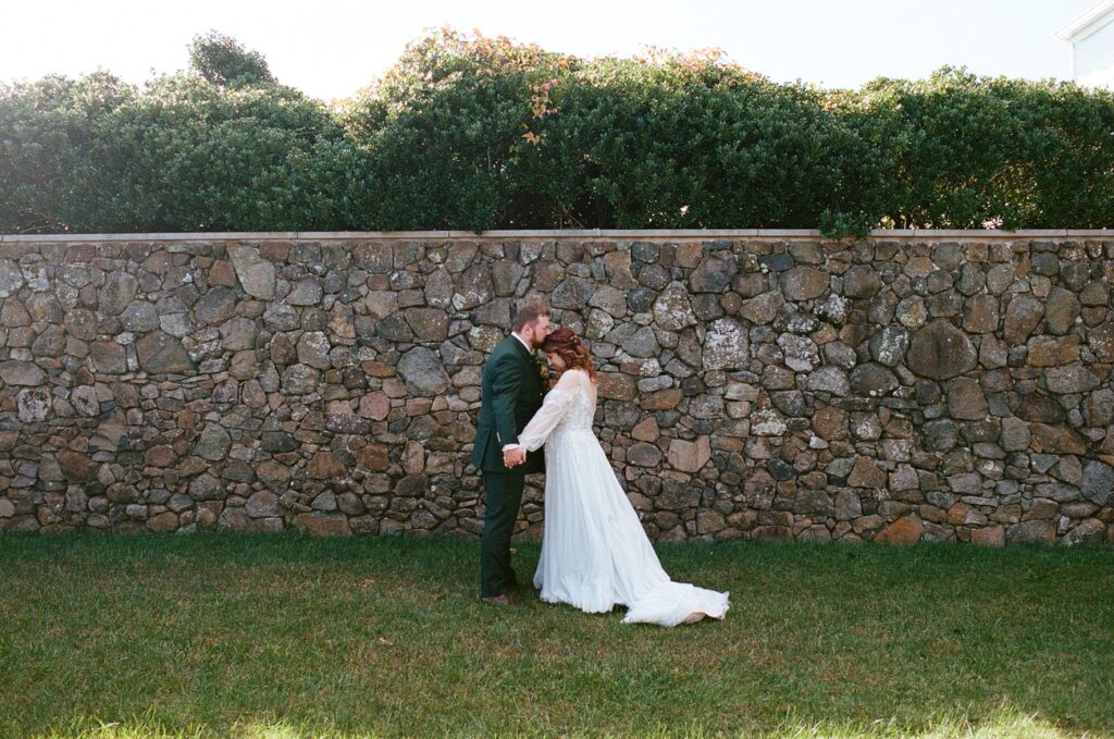 A bride and groom embracing against a stone wall, surrounded by lush greenery, the bride's dress trailing and flowing in the breeze.