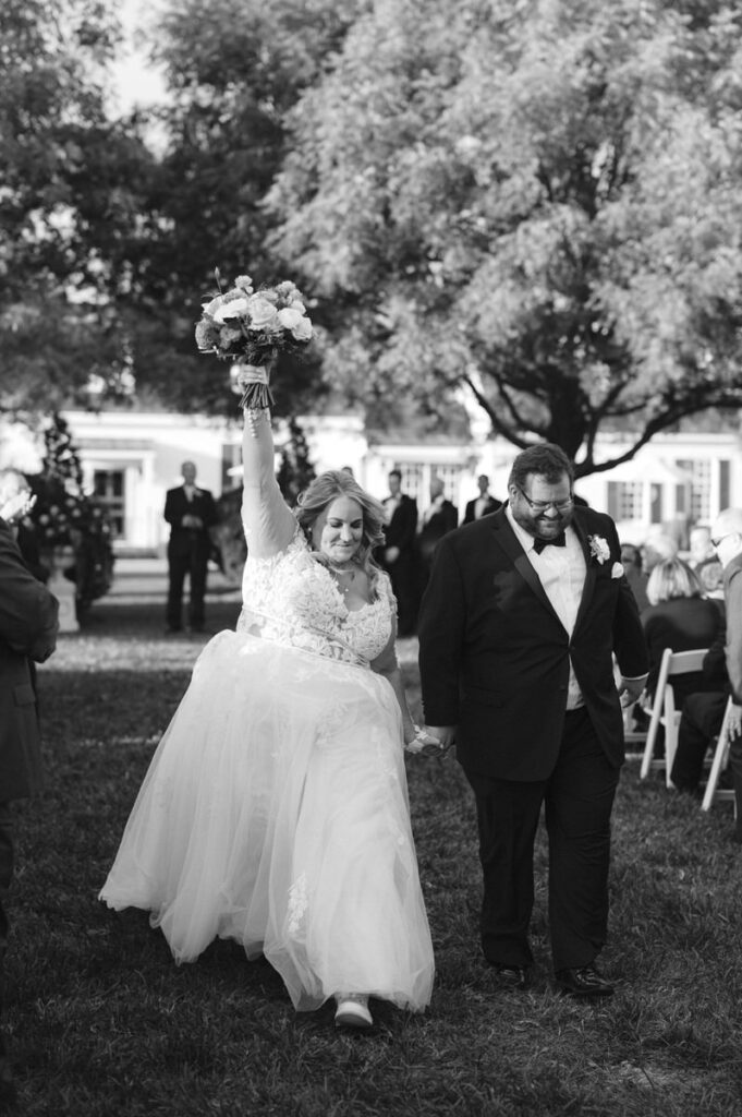 Bride joyfully raises her bouquet in the air while walking down the aisle with her groom, in a black-and-white photo capturing the celebratory spirit post-ceremony