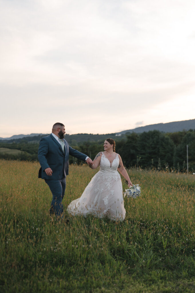 Couple holding hands and walking through a lush meadow at sunset, with the Blue Ridge Mountains in the backdrop, reflecting a serene post-ceremony moment