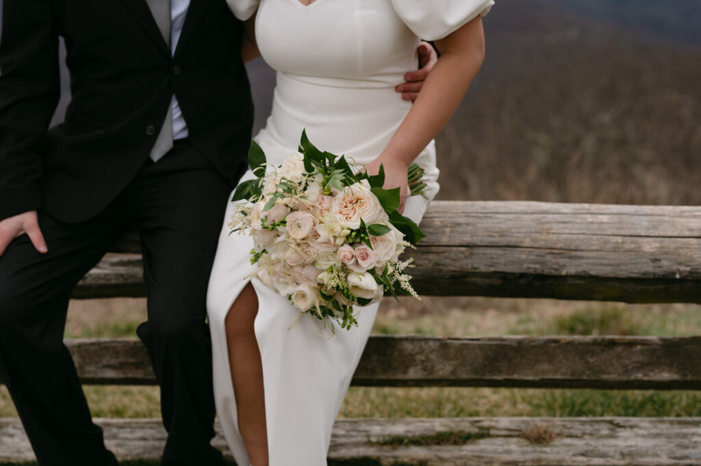 Close-up of a bride and groom at their mountain wedding venue, with the bride holding a delicate white and pink floral bouquet, complementing her elegant white dress