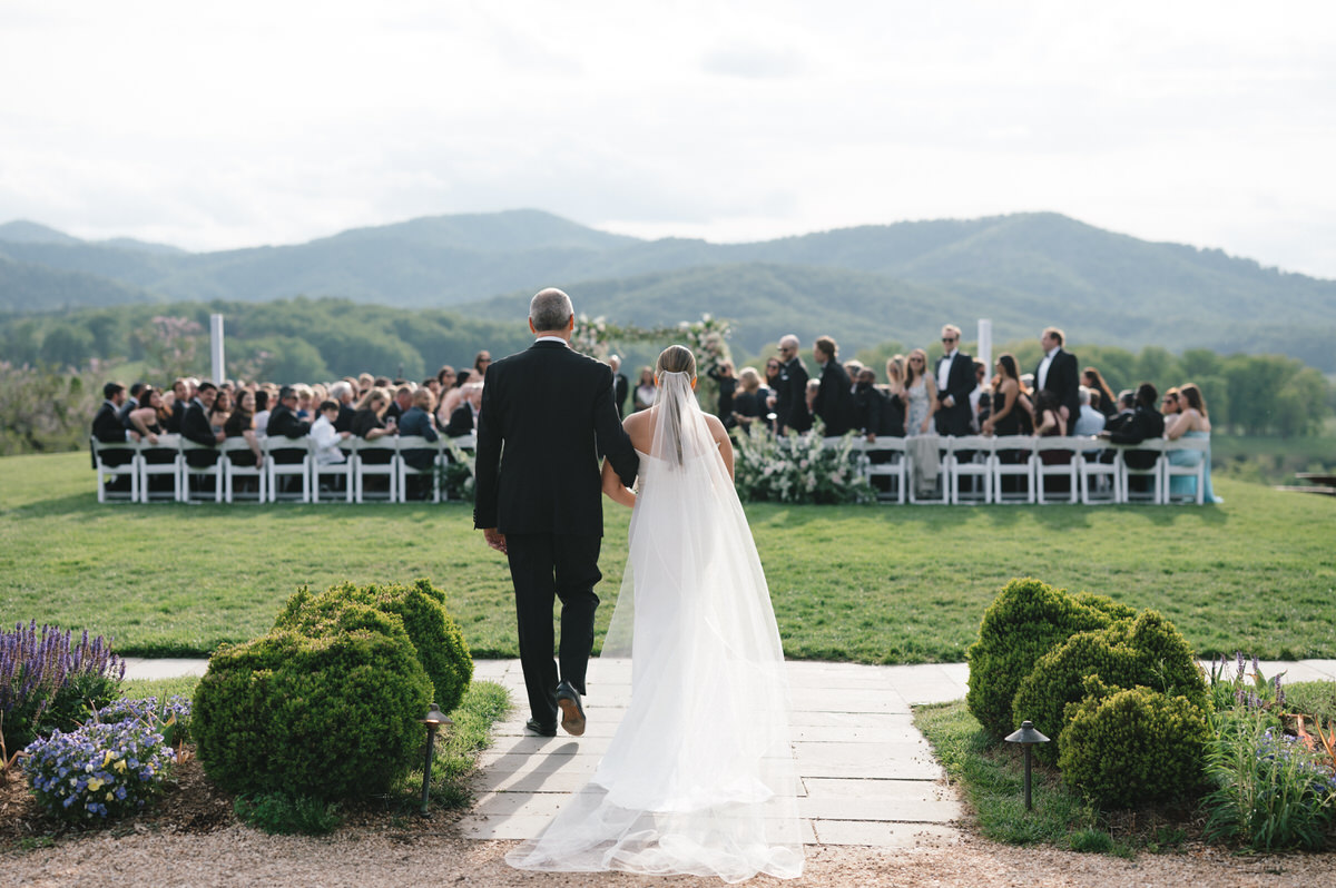 Bride and father walking down the aisle for the ceremony, with guests lined up on either side, and the picturesque Blue Ridge Mountains visible in the distance, creating a stunning backdrop for the wedding venue