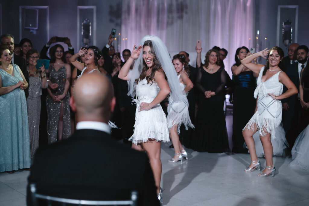 A vibrant wedding reception scene where a bride in a white fringed dress dances exuberantly in front of cheering guests, her joy contagiously spreading throughout the room.