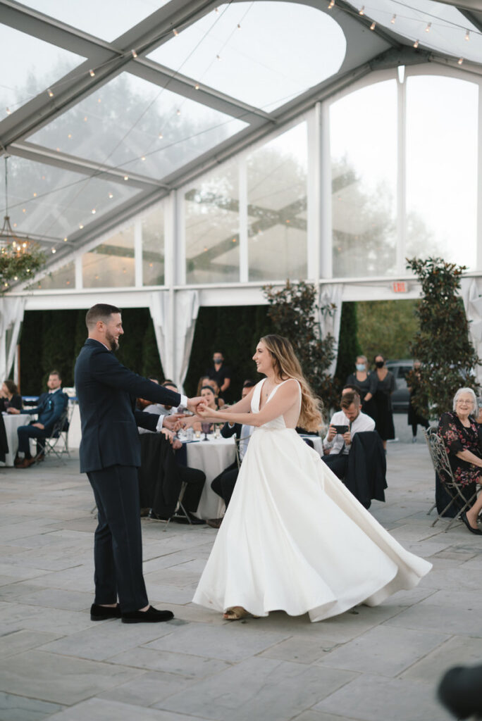 A couple dances under a translucent tented pavilion with string lights, their first dance as newlyweds, with the bride's dress swirling in motion and guests looking on admiringly.