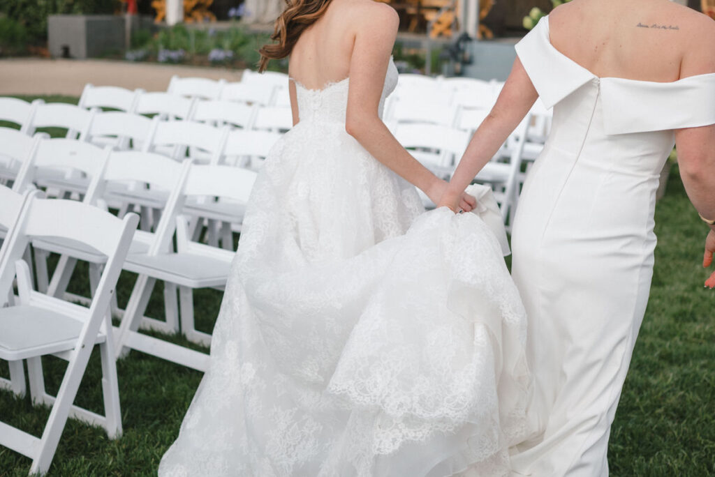 A close-up of two brides from the back, one adjusting the other's lace wedding dress near a row of white ceremony chairs, showcasing the elegance of the gowns and the anticipation of the event.