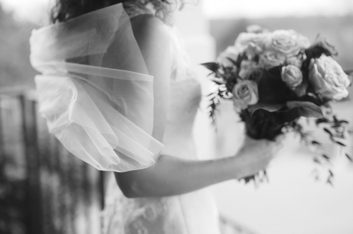 A black and white photo capturing a delicate moment of a bride, her veil fluttering, holding a bouquet of roses, embodying a blend of softness and strength for help deciding between a small wedding and a big wedding