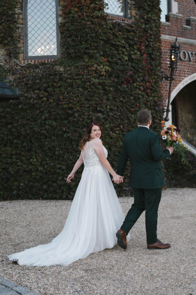 A newlywed couple holding hands and walking together while one looks over their shoulder and smiles. 