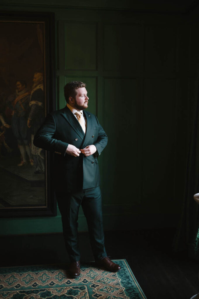 The groom poses by a green wall, looking out contemplatively, dressed in a green suit and adjusting his tie.