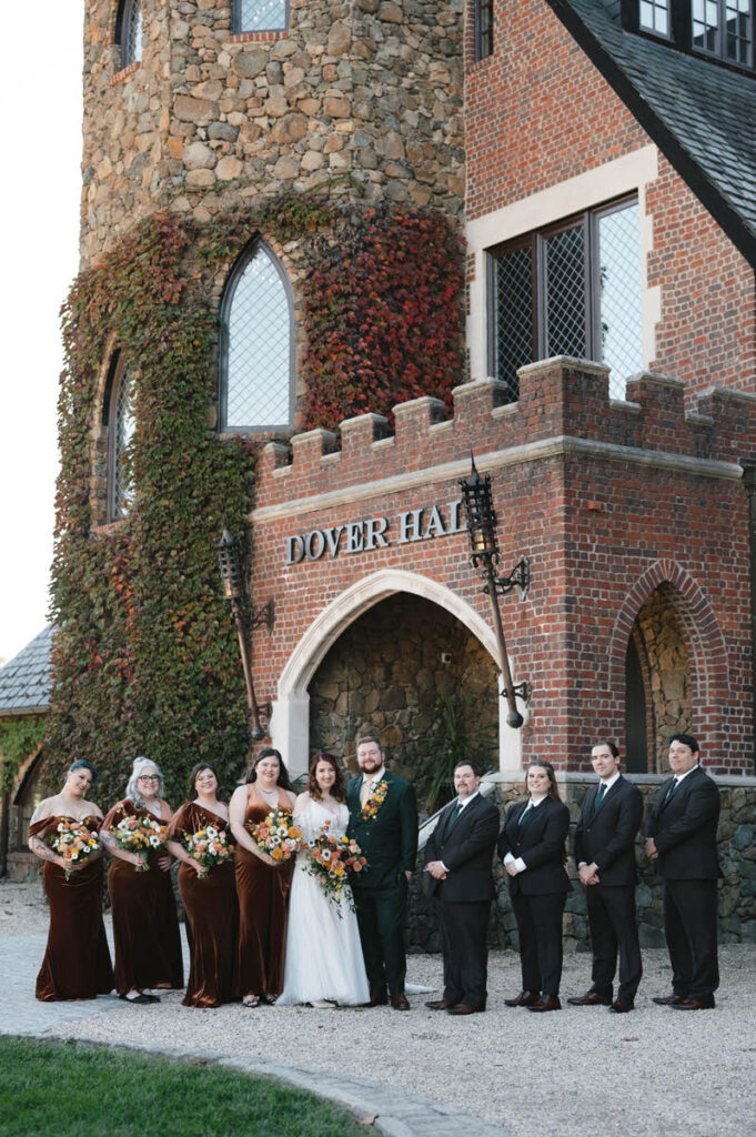 A group photo in front of a castle-like building, featuring the wedding party with bridesmaids in rust dresses and groomsmen in black suits, all smiling.