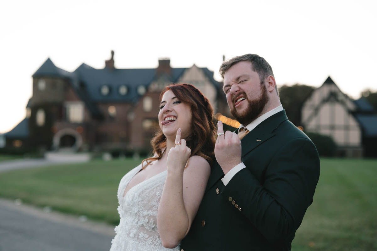 A humorous shot of the bride and groom making playful gestures with their fingers, showing off their new rings, in front of an elegant mansion at dusk.
