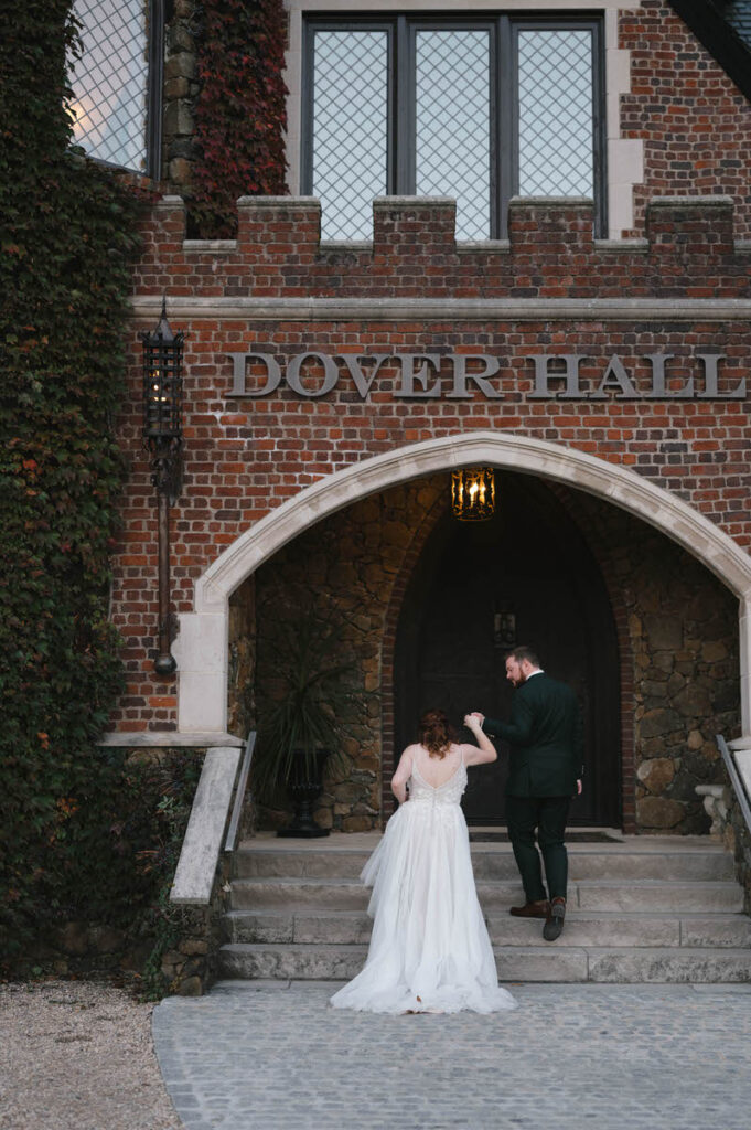 A bride playfully leads her groom up the stone steps of a historic building named "Dover Hall," under a vine-covered archway.