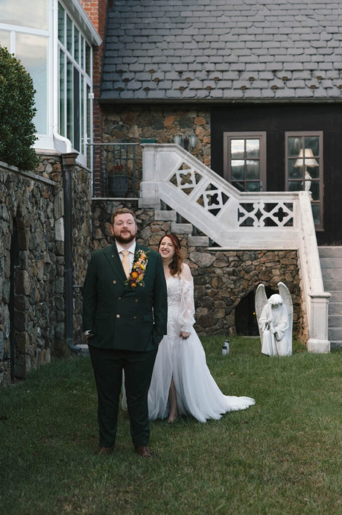 The couple poses smiling on a lush lawn with a dark stone and white wooden building in the background; the groom in a green suit and the bride in a flowing white dress.
