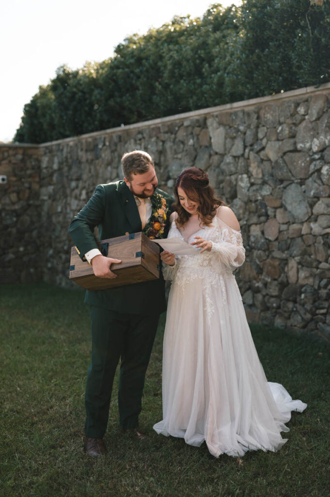 The newlyweds look delighted as they examine the contents of a wooden box together in an open field, dressed in wedding attire.