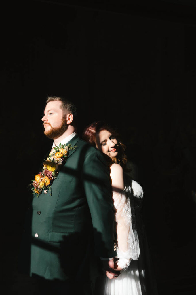 A striking image of the couple standing in a beam of sunlight against a dark background, with the groom in a green suit and the bride leaning on him