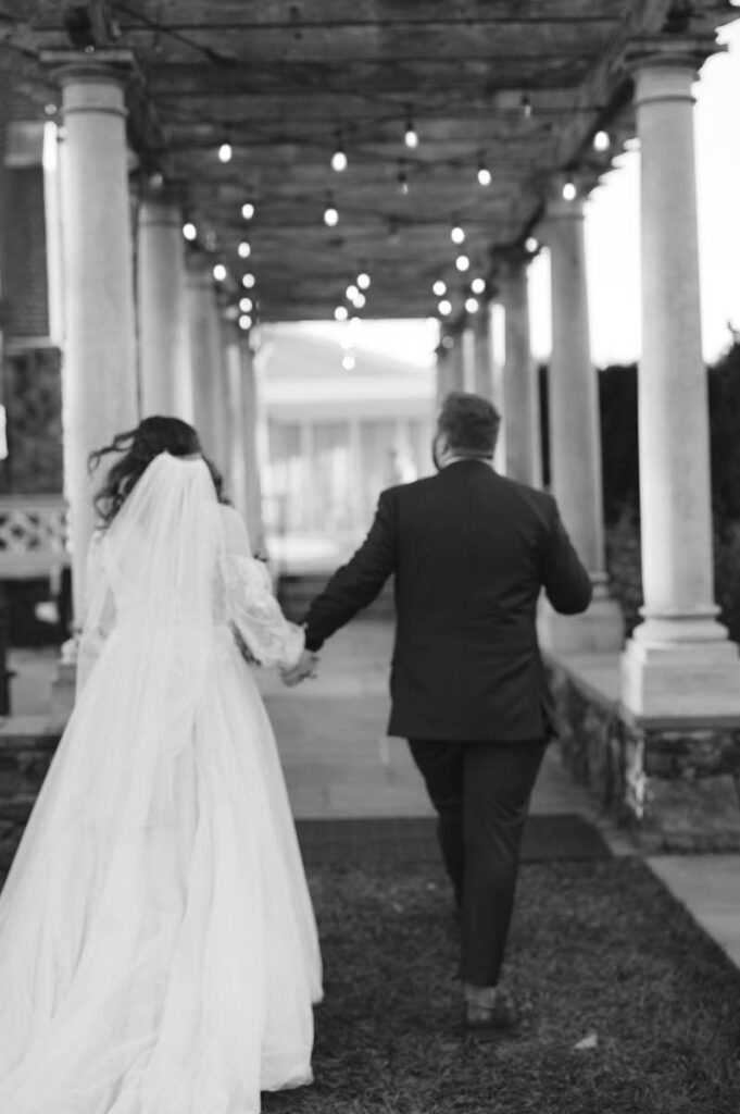 A black and white photo shows the bride and groom walking away hand-in-hand under a pergola adorned with string lights.