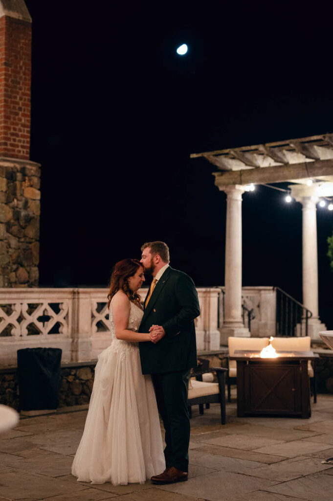 A romantic night scene showing a bride and groom embracing under a moonlit sky, next to a classical architectural colonnade with a glowing fire pit nearby.