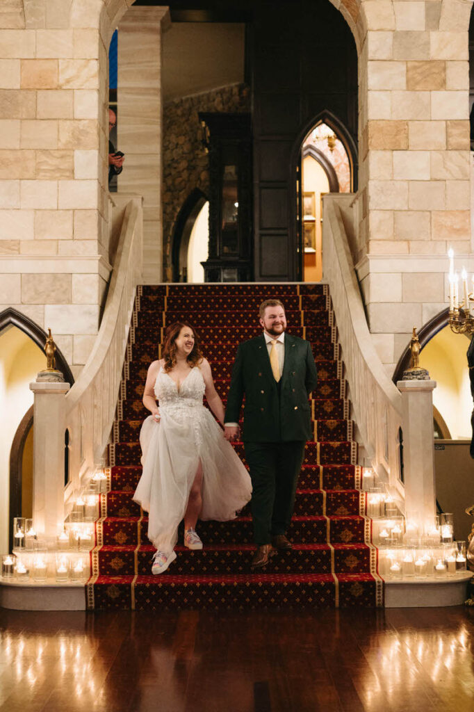 A cheerful bride and groom descending a grand staircase, surrounded by candles, inside an opulent hall with richly patterned carpet and archways.
