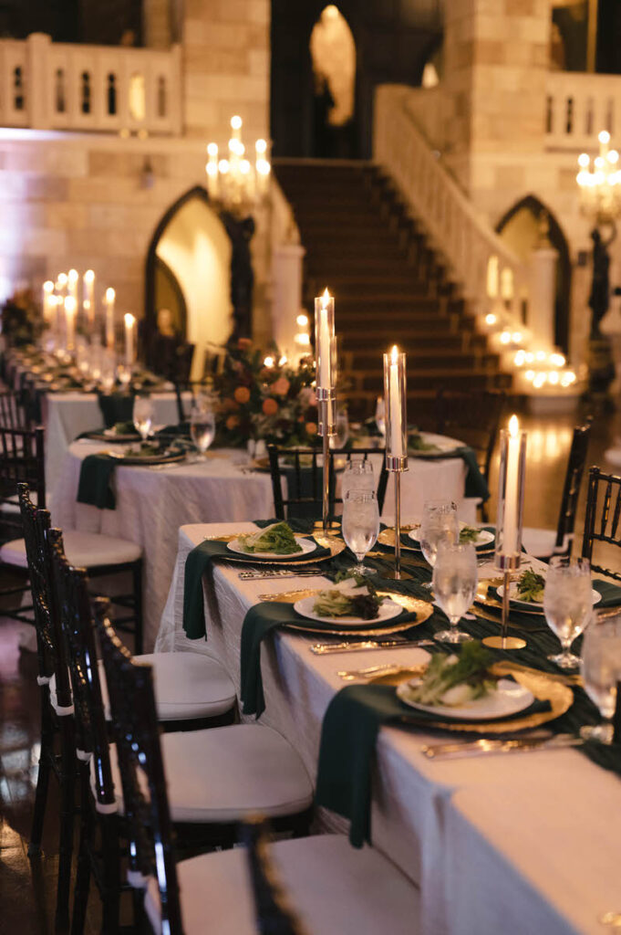 An elegant wedding reception setup inside a grand hall with long dining tables adorned with white linens, green runners, tall candlesticks, and a background of stairs and archways illuminated by soft lighting.