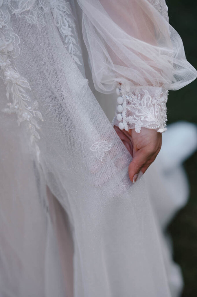 A close-up of a bride's hand gently touching her delicate lace and beaded wedding dress sleeve, showing fine details and craftsmanship.