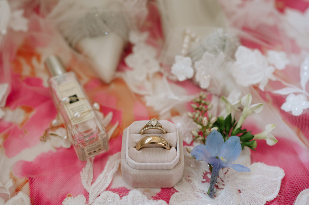 A close-up shot of a wedding arrangement featuring a pair of gold rings in a white velvet box, a bottle of Jo Malone perfume adorned with small pearls, and delicate flowers on a vibrant pink floral fabric.