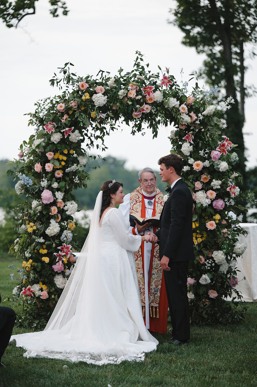 A bride and groom stand before a floral arch, holding hands and exchanging vows during their outdoor wedding ceremony, with a priest officiating the service. The arch is adorned with an abundance of colorful flowers, including roses and lilies, set against a lush, green backdrop.