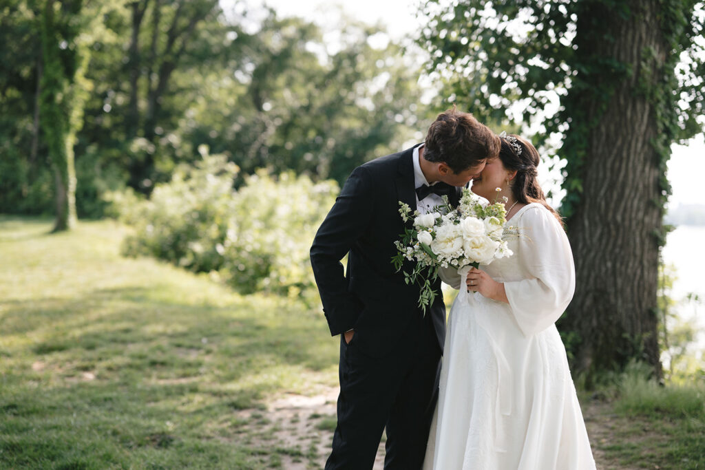 A bride and groom share a kiss outdoors, surrounded by lush greenery. The bride holds a bouquet of white flowers and greenery, while the groom, dressed in a black suit, leans in for the kiss.